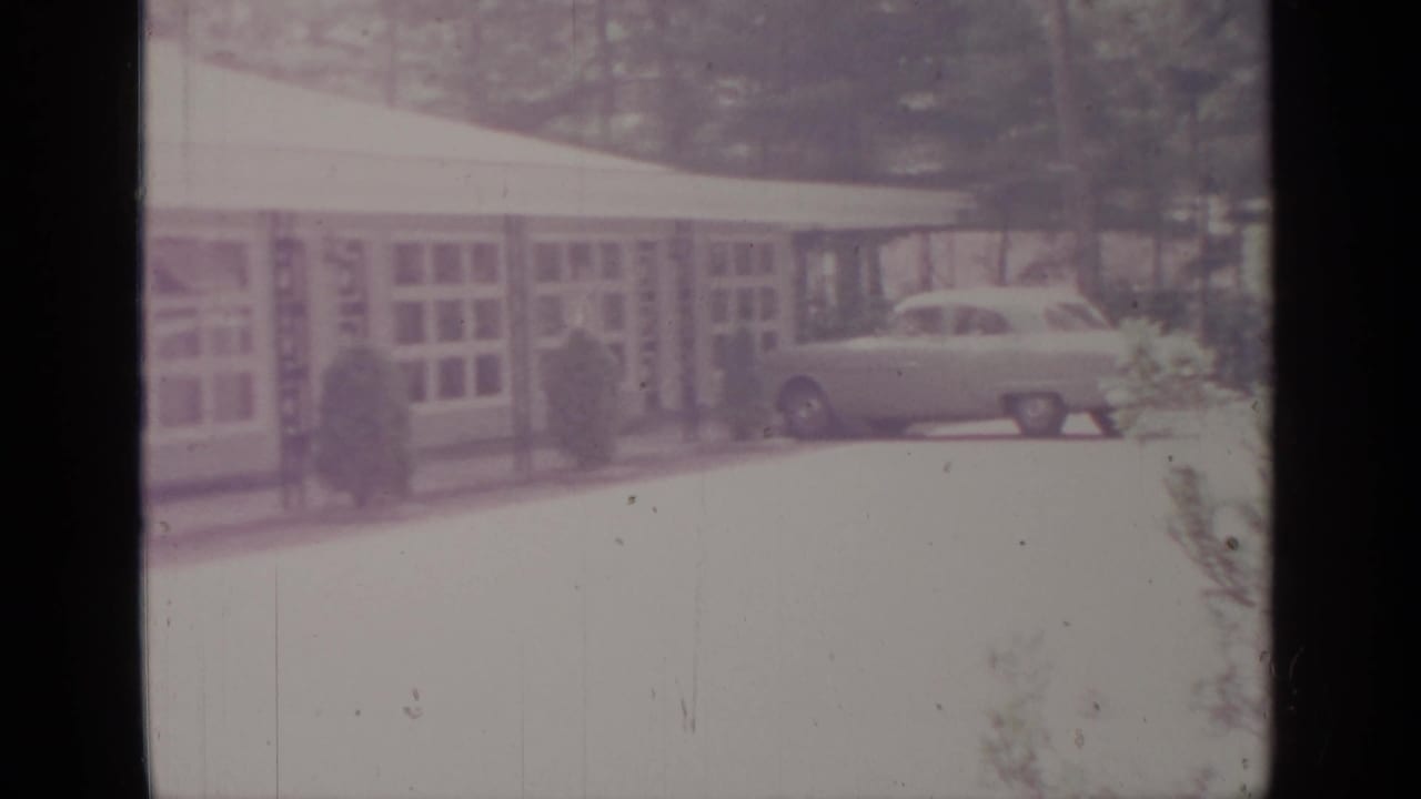 1964 Greenwood Lake exterior view of a low windowed building with shrubs and a light-colored car parked at the edge of the frame.