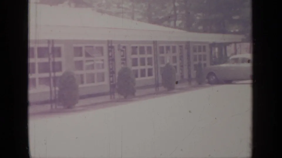 1964 Greenwood Lake frame showing the building's windowed exterior, trimmed shrubs, pavement, and the front portion of a parked car.