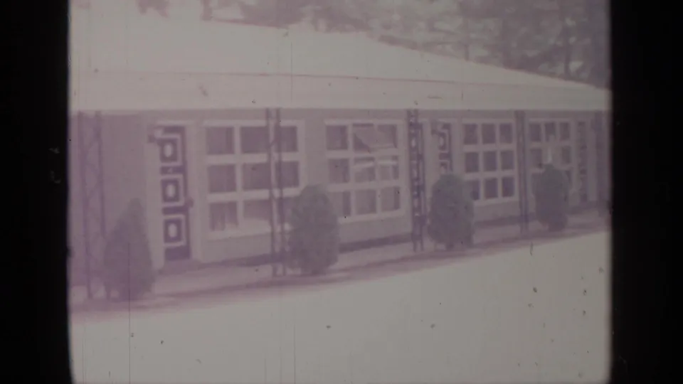 1964 Greenwood Lake 8mm frame centered on a low building facade with large windows, geometric door panels, and small shrubs.