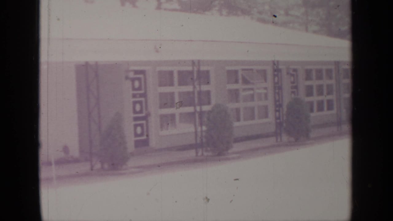 1964 8mm frame from Greenwood Lake showing a low mid-century building facade with large windows, geometric door panels, shrubs, and a pale foreground.
