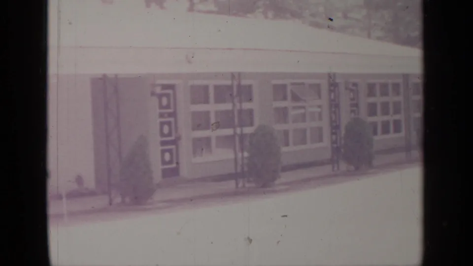 1964 8mm frame from Greenwood Lake showing a low mid-century building facade with large windows, geometric door panels, shrubs, and a pale foreground.