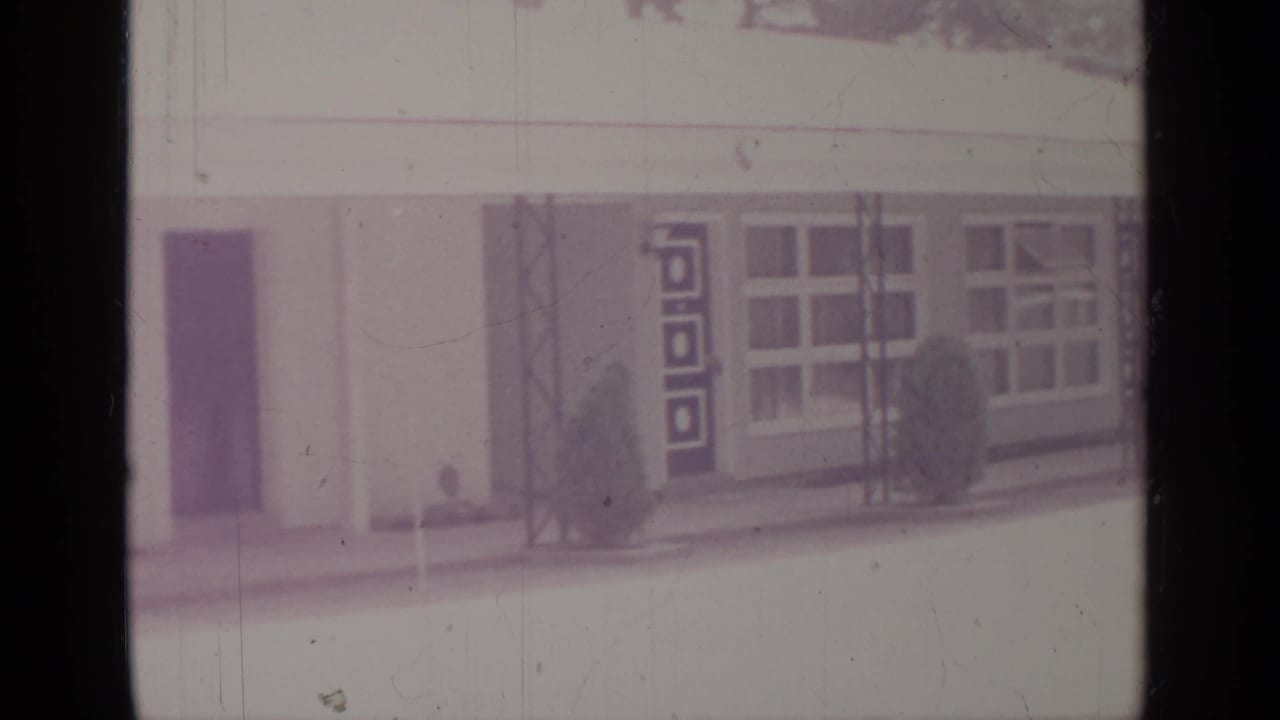 Greenwood Lake 1964 exterior frame showing a dark doorway, decorative geometric panel, broad windows, metal posts, and small shrubs along the facade.