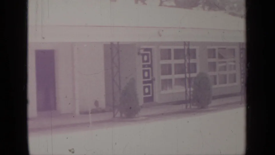 Greenwood Lake 1964 exterior frame showing a dark doorway, decorative geometric panel, broad windows, metal posts, and small shrubs along the facade.
