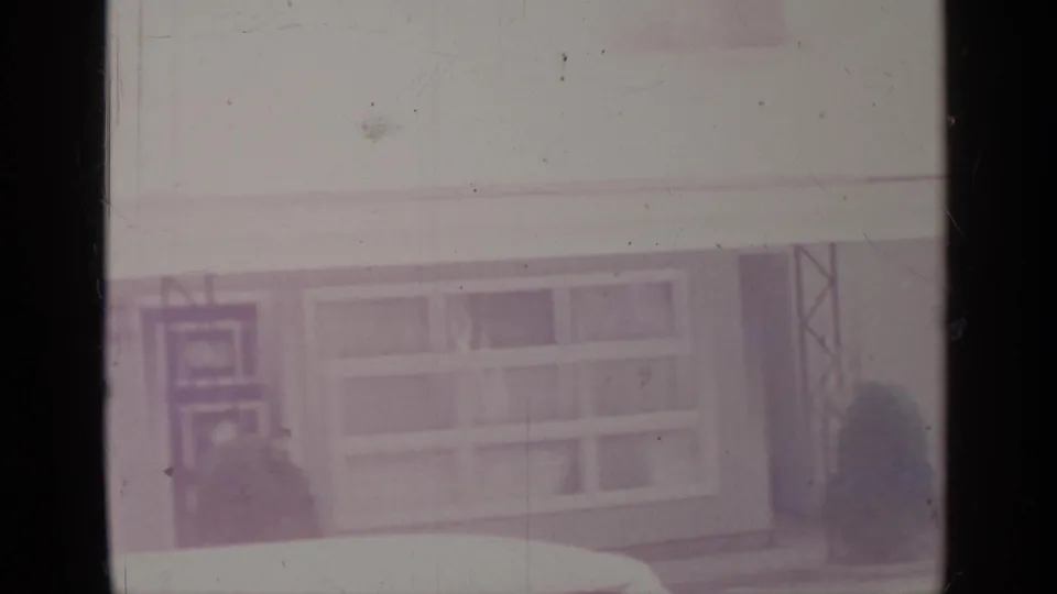 Soft 8mm frame showing the building facade closer, with window bays, part of a patterned door, small shrubs, vertical posts, and a bright foreground band.