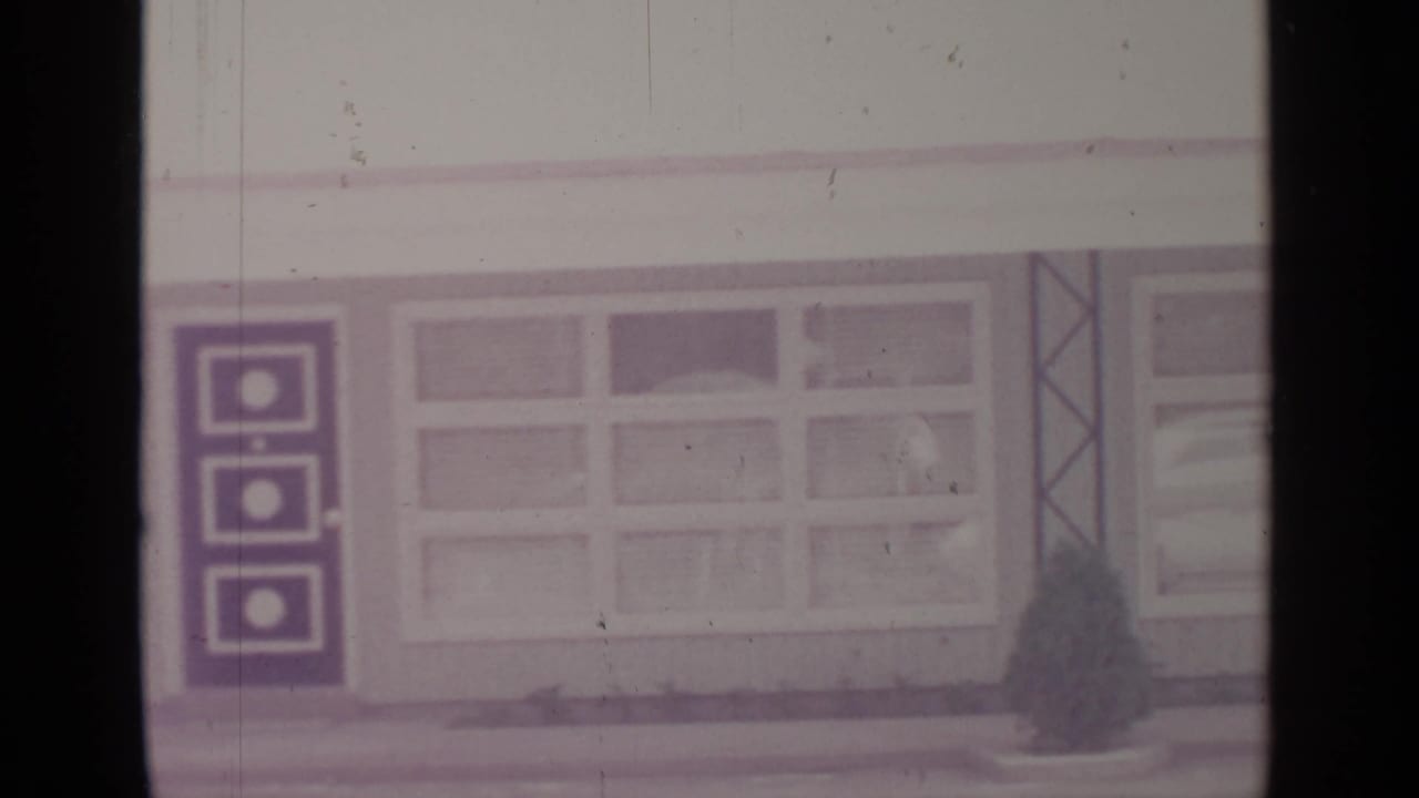 Faded 8mm frame of a Greenwood Lake street-side building with large windows and a dark panel, likely from the 1964 home movie.
