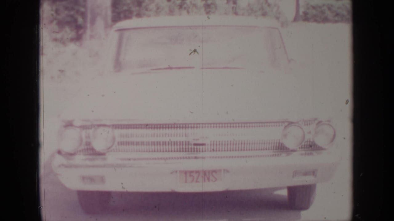 A vintage American car faces the camera on a quiet Greenwood Lake street in a faded 1964 8mm frame.