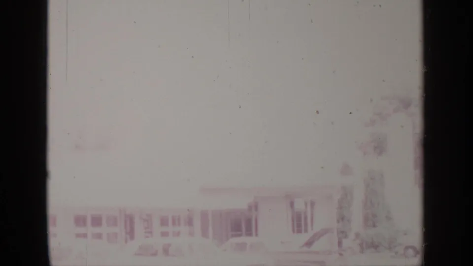 A bright, washed 8mm frame shows a Greenwood Lake building and portions of parked vehicles along the street.