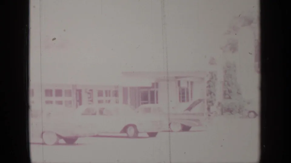 Faded 1964 8mm footage shows parked cars along a Greenwood Lake street beside a low building and trees.