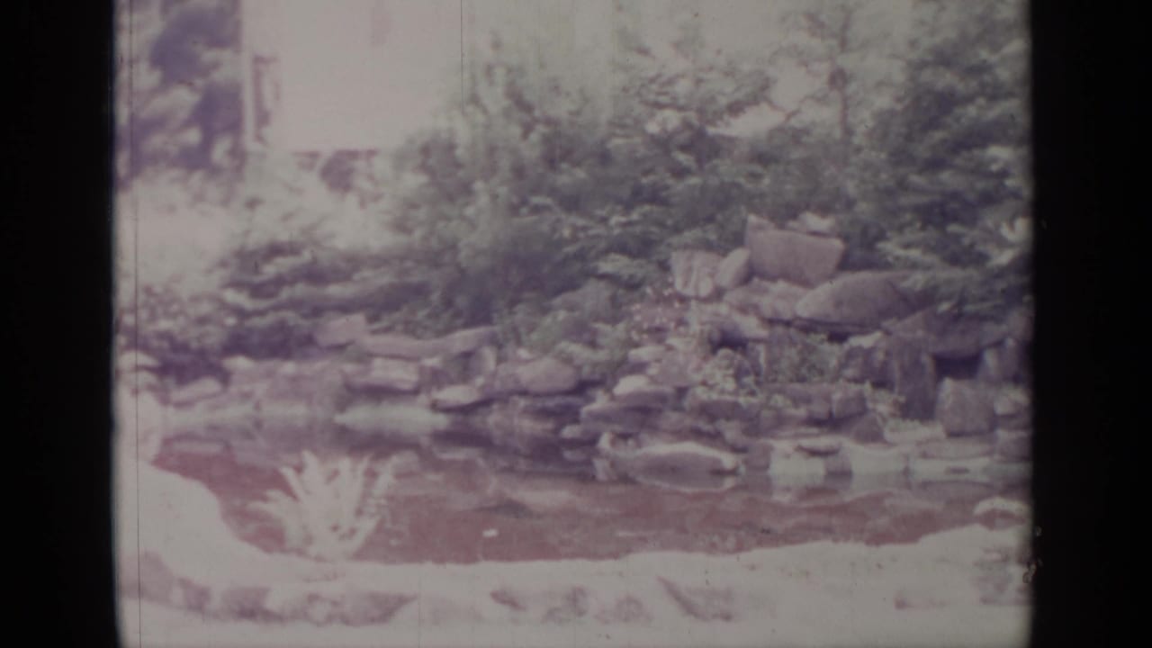 1964 Greenwood Lake frame with a rock water feature, pond surface, shrubs, and bright overexposed garden edges.