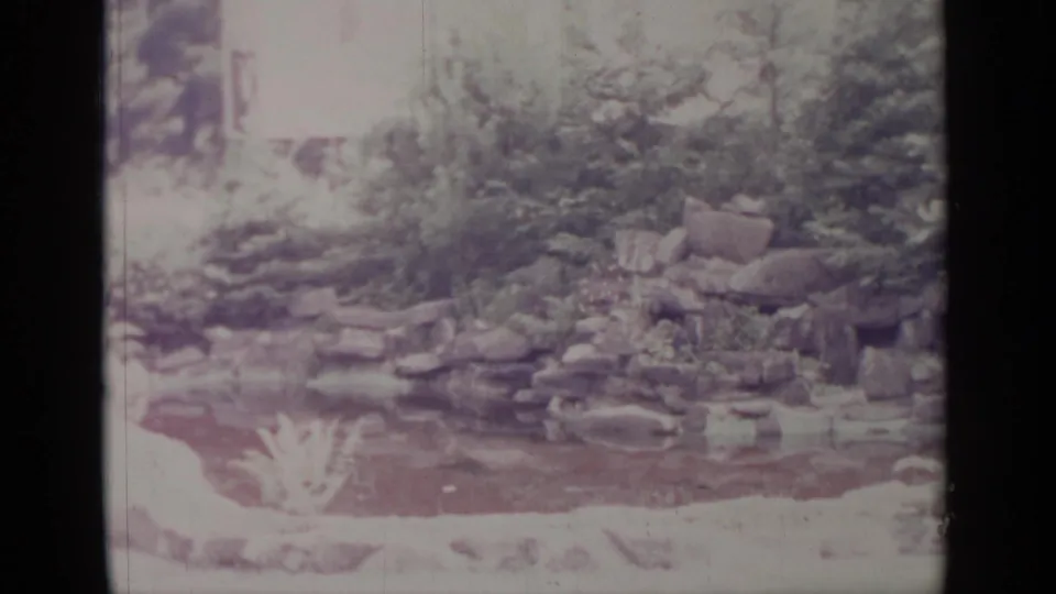 1964 Greenwood Lake frame with a rock water feature, pond surface, shrubs, and bright overexposed garden edges.