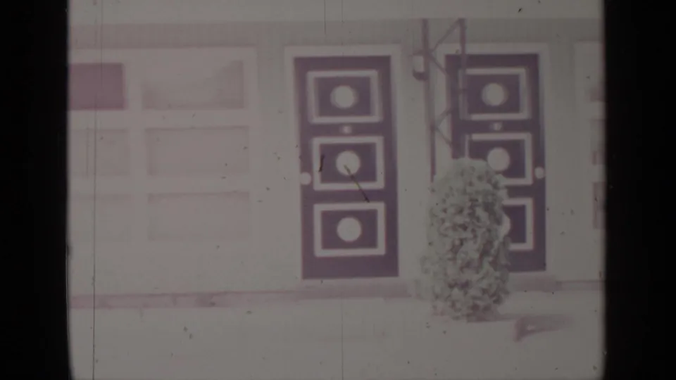 1964 Greenwood Lake frame showing a snowbound building exterior with dark geometric door panels, windows, and shrubs.
