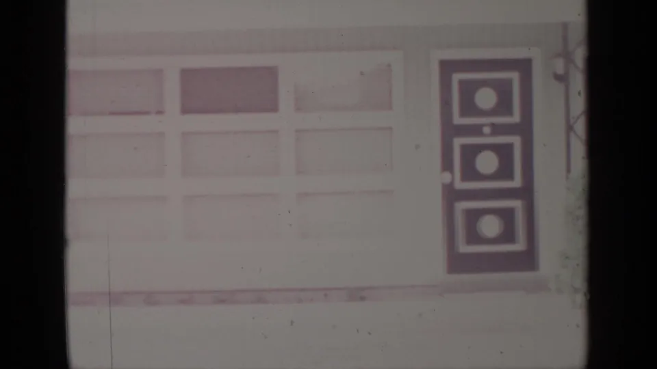 1964 Greenwood Lake home-movie frame showing a pale building facade, garage-like panels, and a dark geometric door in winter snow.
