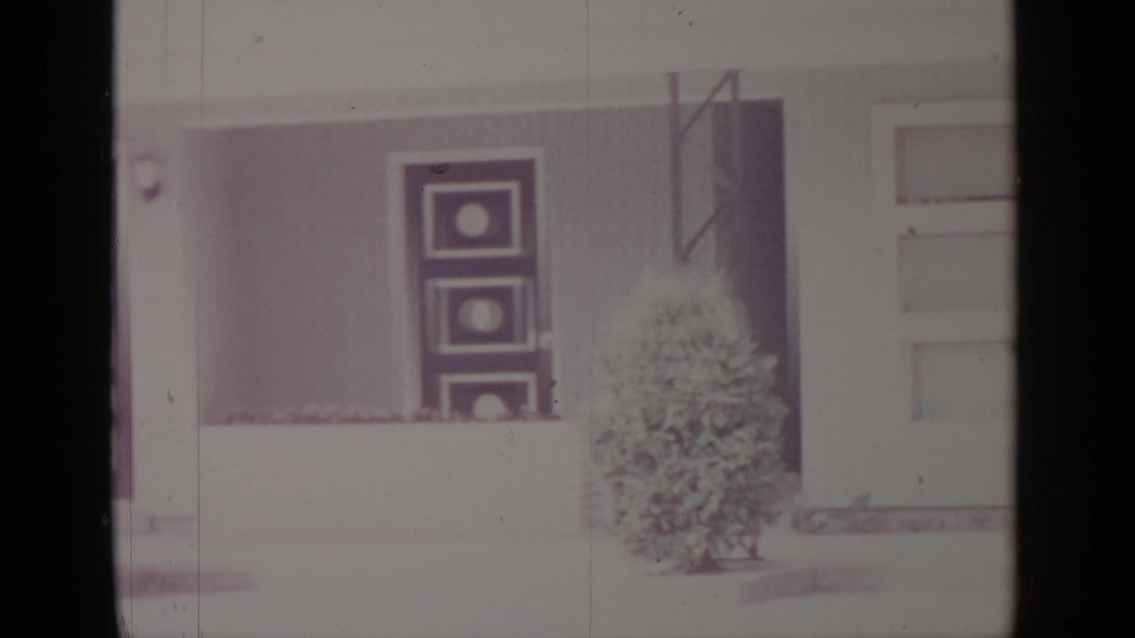 White-sided house exterior with a front door, window boxes, shutters, and a small evergreen shrub.