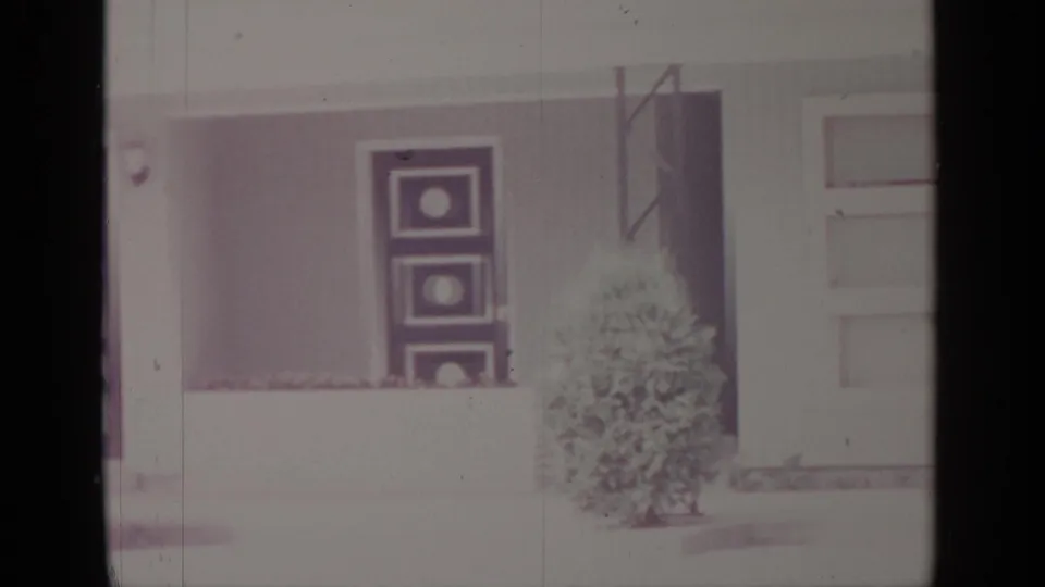 1964 Greenwood Lake home-movie frame showing a white house exterior with a front door, dark shutters, flower boxes, and a small evergreen shrub.