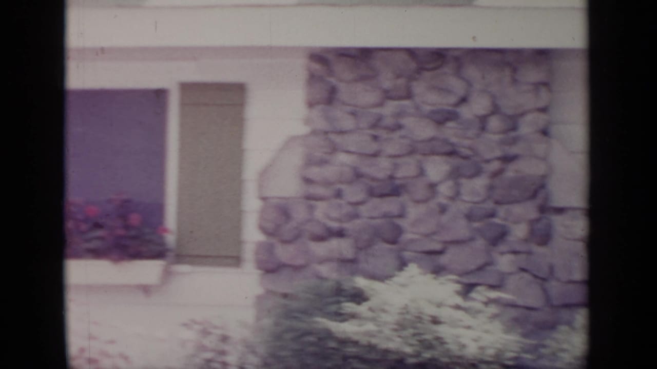 1964 Greenwood Lake frame showing a stone wall section, shuttered window, flower box, and garden plantings on a house exterior.