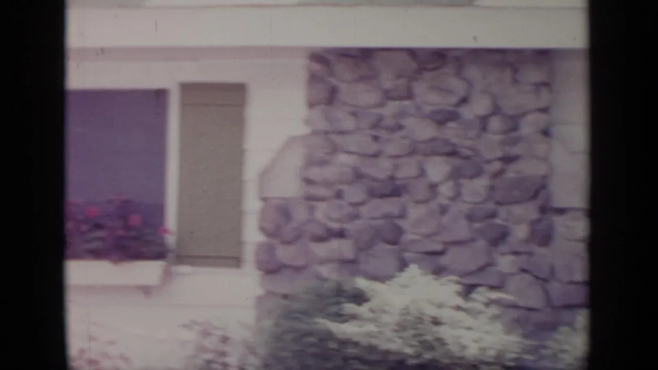 1964 Greenwood Lake frame showing a stone wall section, shuttered window, flower box, and garden plantings on a house exterior.