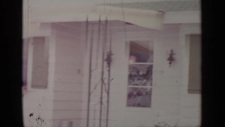 1964 Greenwood Lake home-movie frame showing part of the house exterior with shutters, a wall ornament, and tall vertical rods near the wall.