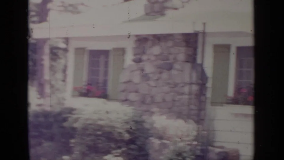 1964 Greenwood Lake frame showing a stone exterior section between shuttered windows, with flower boxes and shrubs along the house.