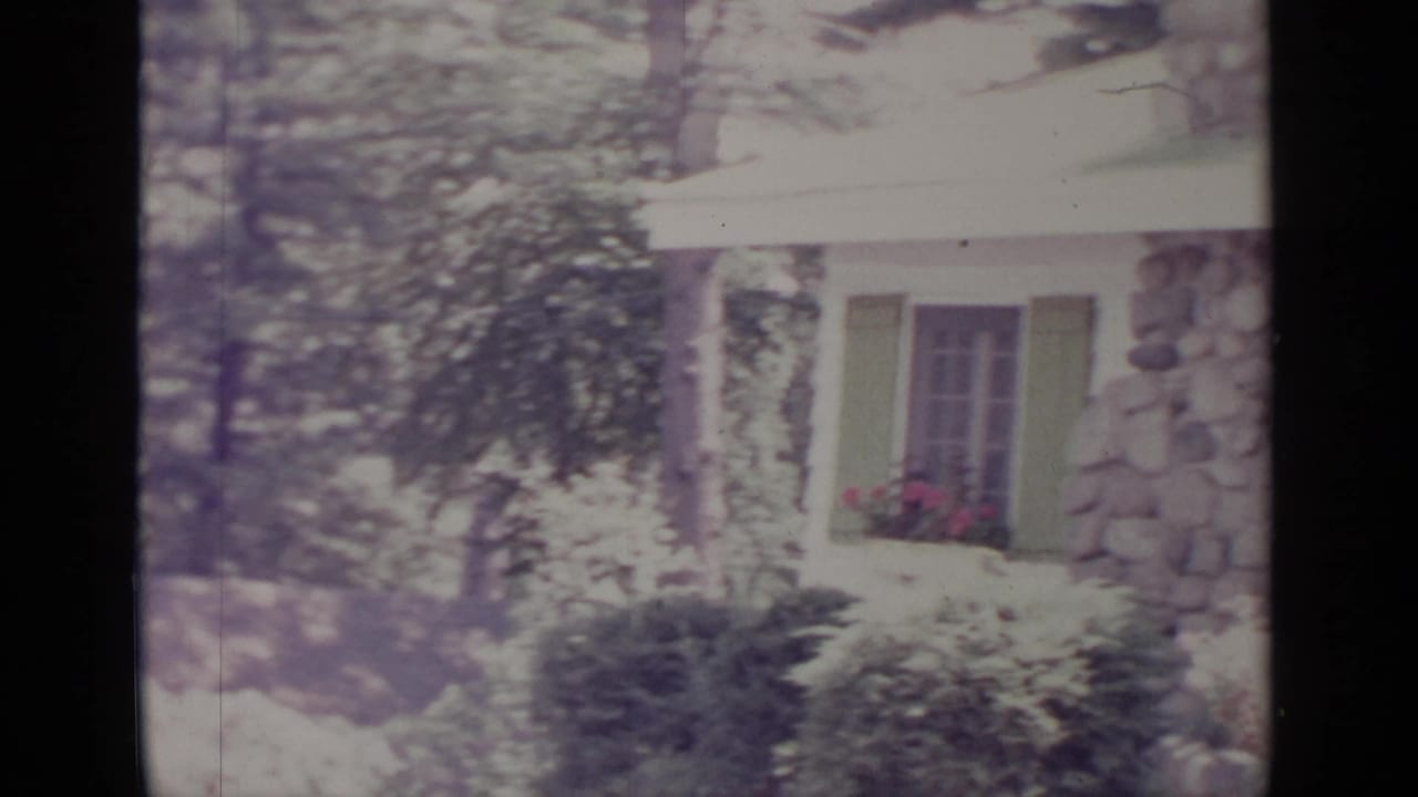 White house with green shutters, stone facade, shrubs, trees, and flower box.