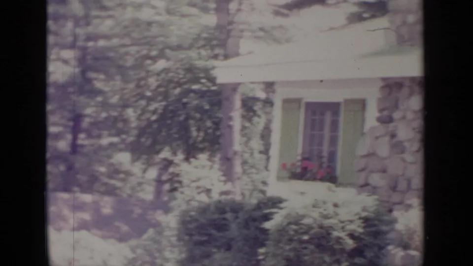 1964 Greenwood Lake home-movie frame showing a white-sided house with green shutters, stonework, trees, shrubs, and pink flowers.
