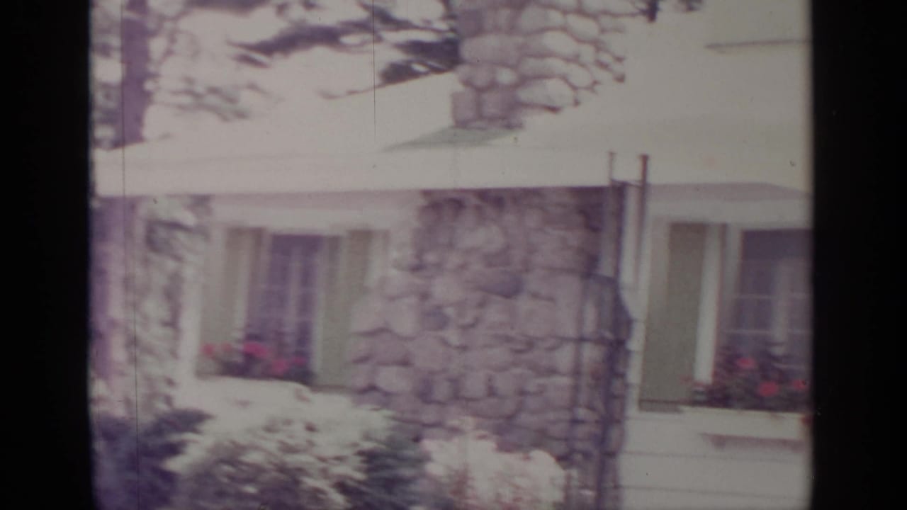 1964 Greenwood Lake frame of a house exterior with stone facade, green shutters, shaded windows, shrubs, and pink window flowers.