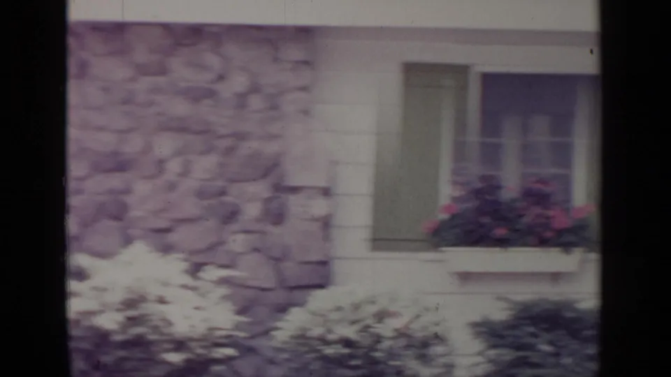 1964 Greenwood Lake frame highlighting a shuttered window, pink flower box, stone house wall, and sunlit shrubs.