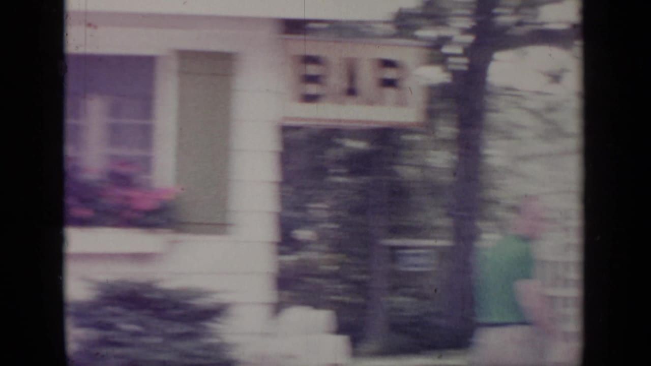 Blurred frame of a man near a white house exterior with dark film borders.