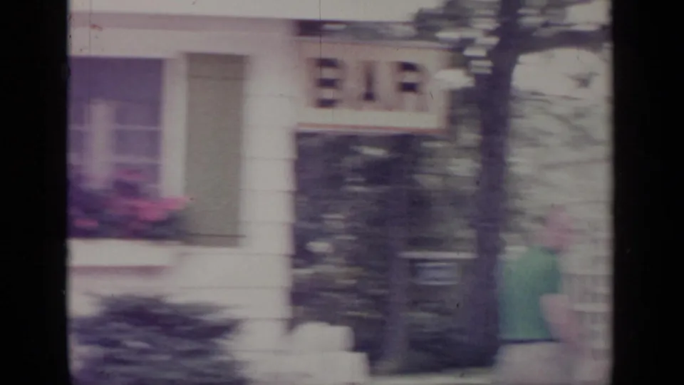 Blurred 1964 Greenwood Lake 8mm frame showing a man moving near a white house exterior and window area.