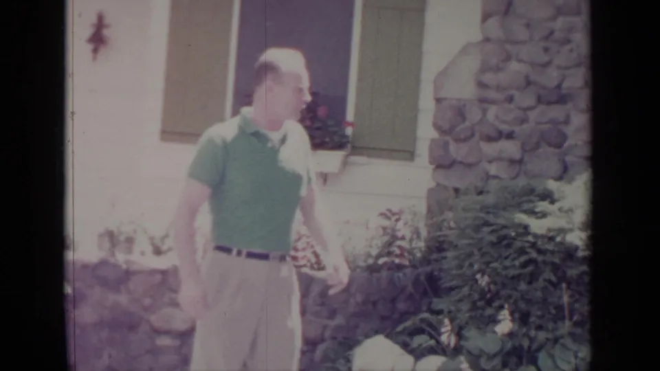 The man moves along the garden outside the Greenwood Lake house, with shrubs, rocks, and stonework behind him.