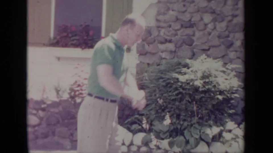 A man in a green polo looks down near shrubs and garden rocks outside the Greenwood Lake house in 1964.