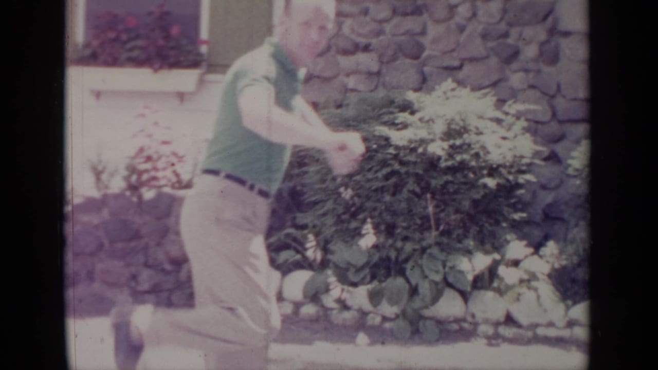 1964 Greenwood Lake home-movie frame showing a man in a green polo and light pants posing in a yard by a stone wall and window box.