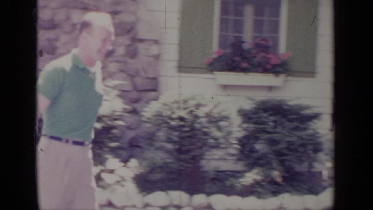 Man in green polo laughing or smiling beside a house wall and shrubs.