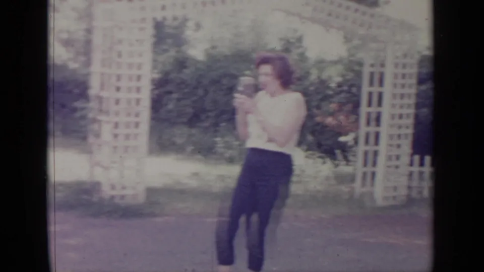 1964 Greenwood Lake home-movie frame showing a woman in a white top and dark pants photographing someone in a yard.