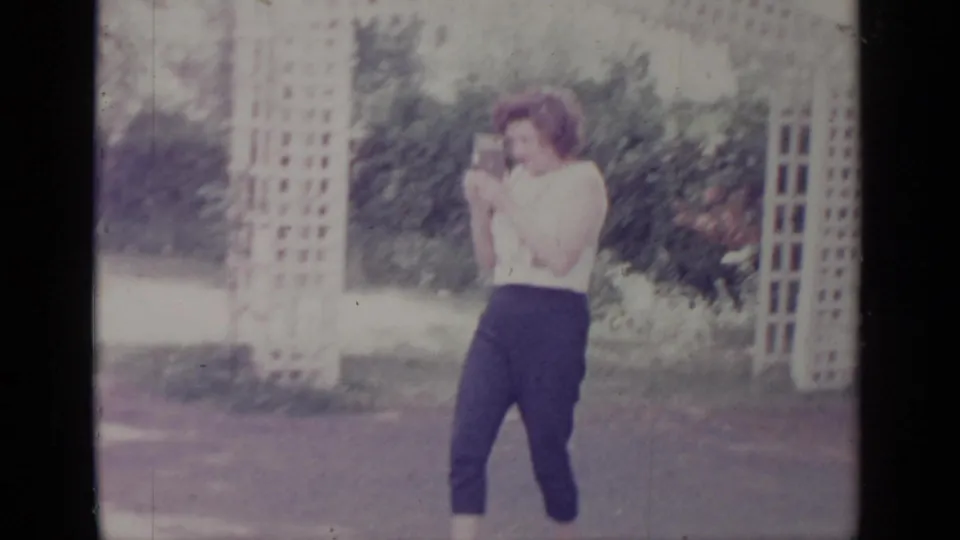 1964 Greenwood Lake frame of a woman holding a camera in a residential yard, with trellises and greenery behind her.