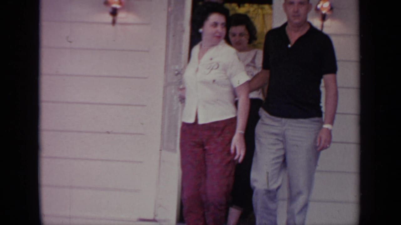 Adults leave a Greenwood Lake house doorway, with a woman in a white blouse and red pants beside a man in a dark polo shirt.