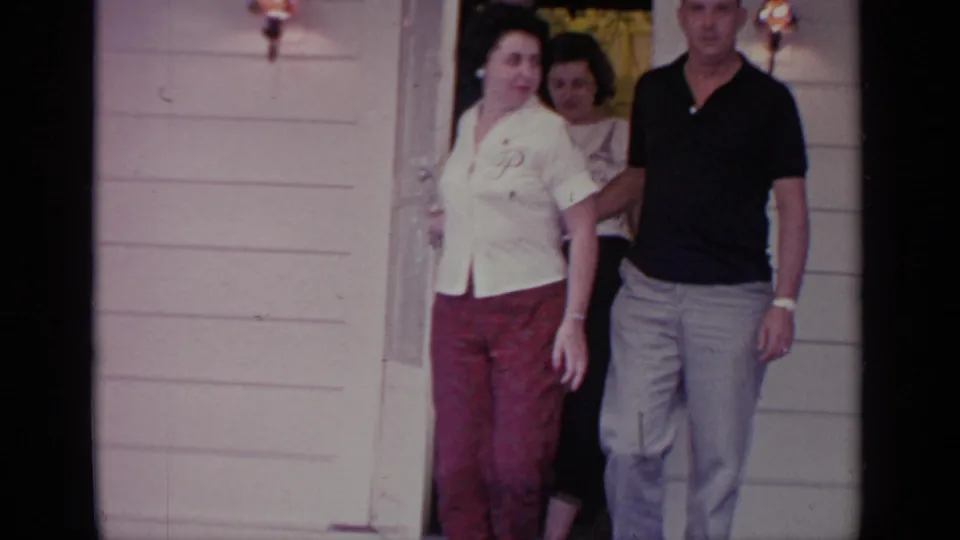 Adults leave a Greenwood Lake house doorway, with a woman in a white blouse and red pants beside a man in a dark polo shirt.