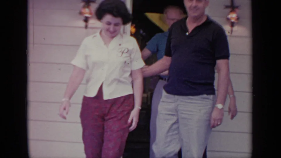 A 1964 home-movie frame shows a woman in a monogrammed white blouse walking from the doorway with a man close behind.
