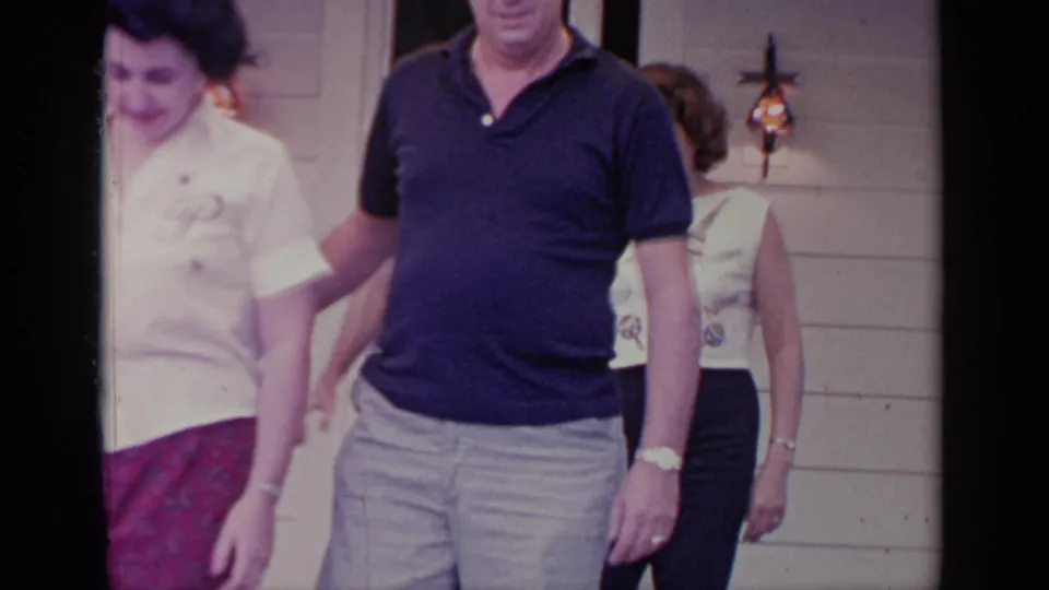 Adults move past the camera outside the Greenwood Lake house, with the doorway and another woman still visible behind them.