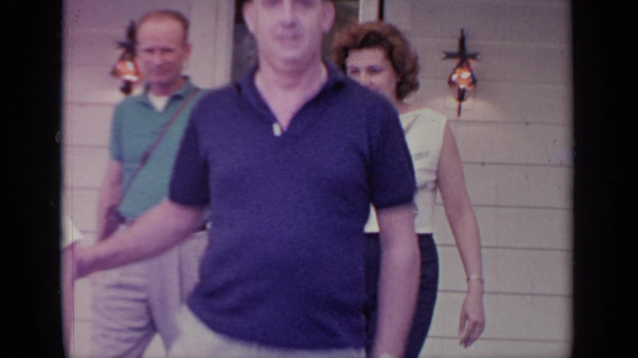 Adults walking past a white-sided house, with a man in a navy polo closest to the camera.