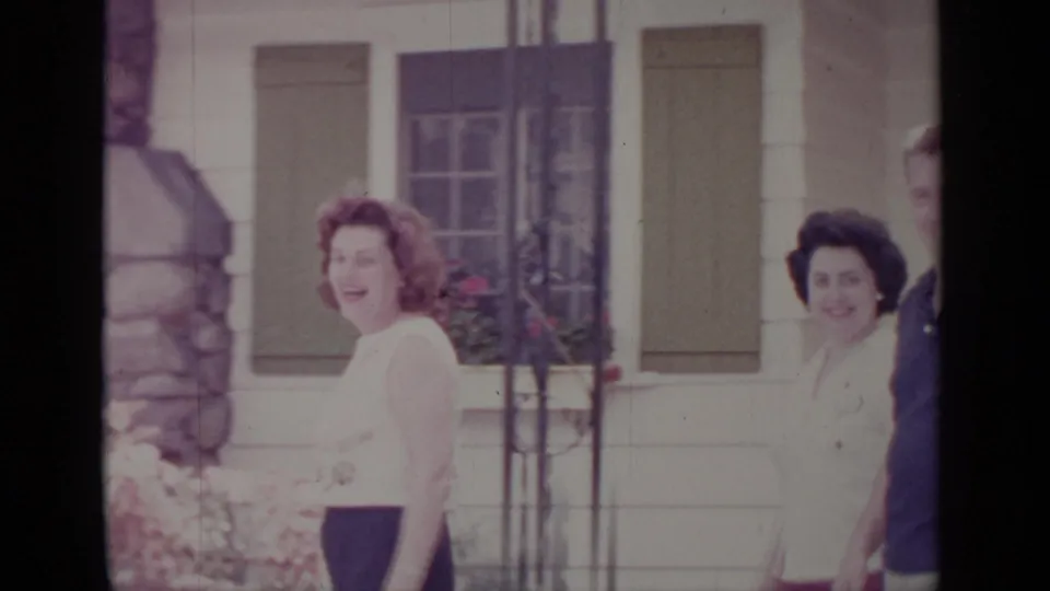 1964 home-movie frame of two women walking near a white-sided Greenwood Lake house with shutters and stonework.