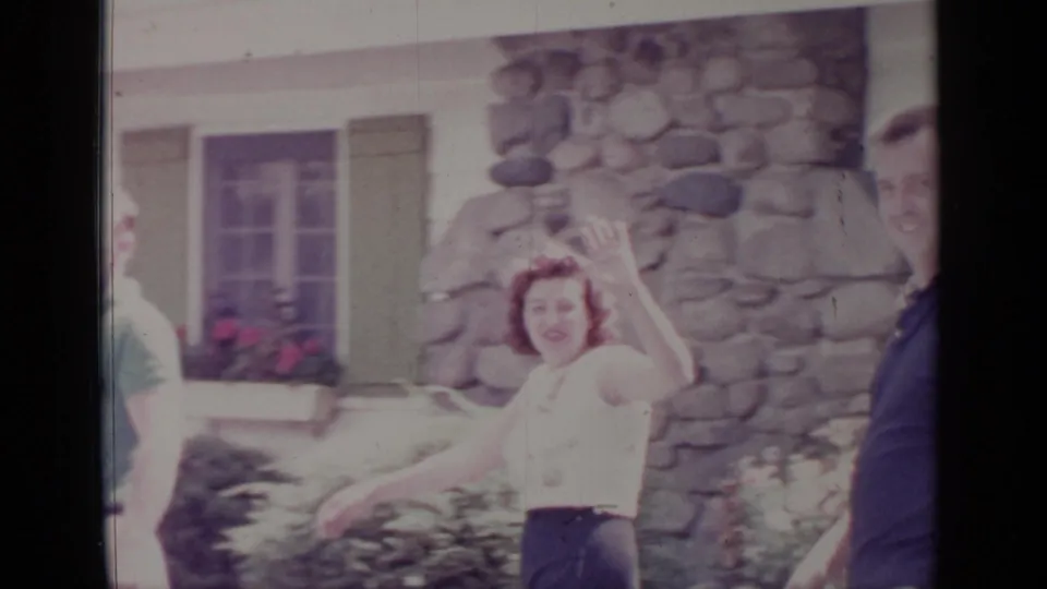 A woman waves to the 8mm camera outside a Greenwood Lake house, with stonework and other adults partly in view.