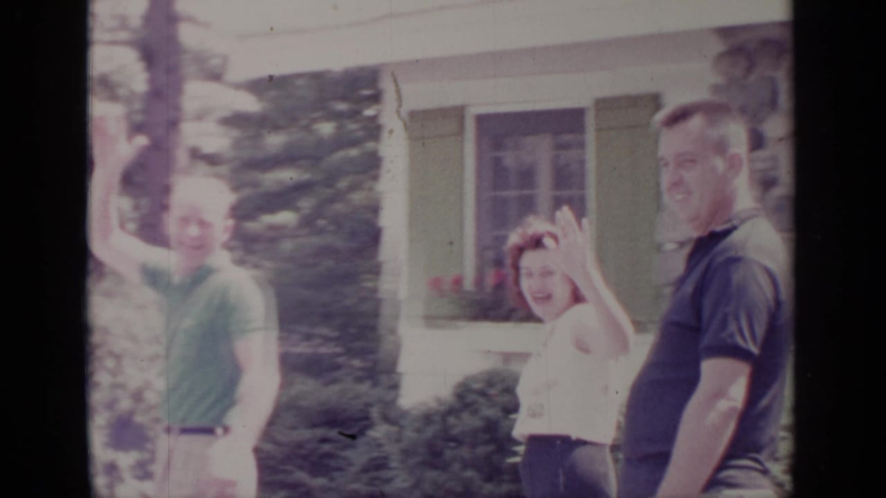 Three adults stand outside a house while two wave toward the camera.