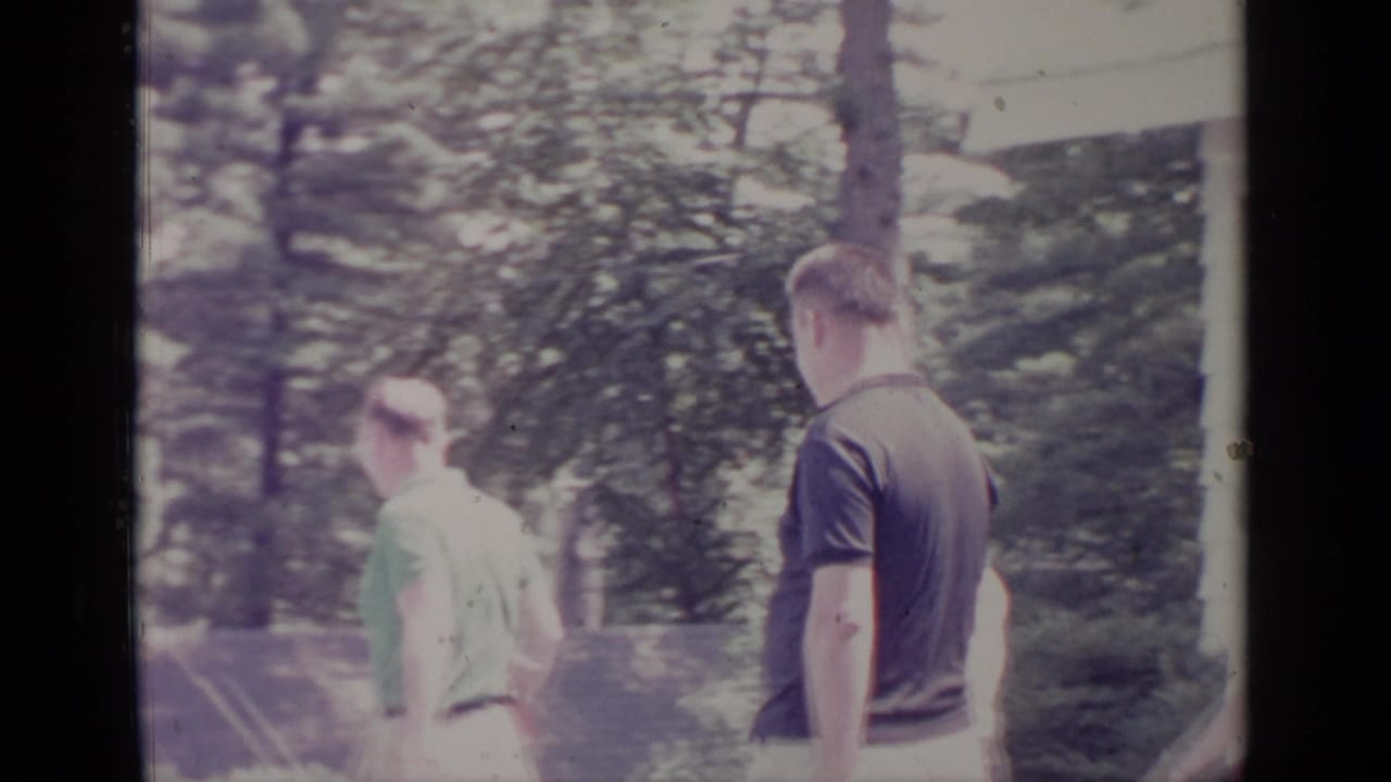 1964 Greenwood Lake frame with two adult men in casual shirts moving through a shaded yard with trees and shrubs.