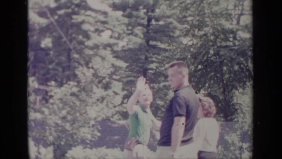 1964 Greenwood Lake home-movie frame showing adults in a yard, one raising both hands while others stand nearby.