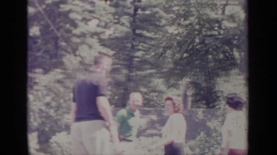 1964 Greenwood Lake frame showing a small group of adults gathered in a leafy yard, one person turned toward the others.