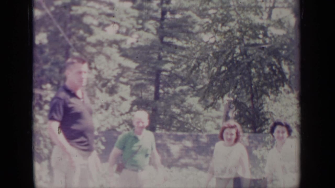 1964 Greenwood Lake home-movie frame showing adults in casual clothing walking through a bright evergreen forest area.