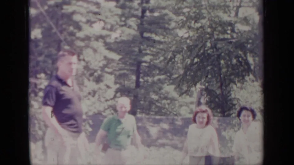 1964 Greenwood Lake home-movie frame showing adults in casual clothing walking through a bright evergreen forest area.