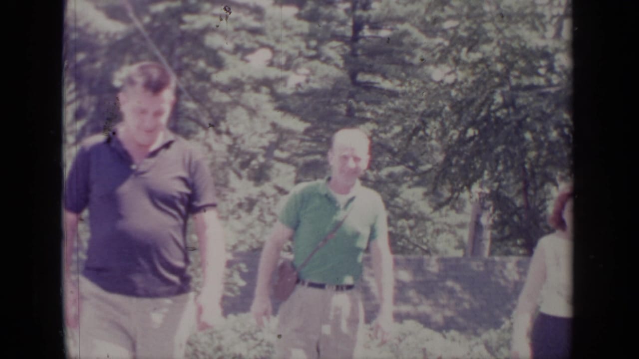 1964 Greenwood Lake frame showing two casually dressed men walking near evergreens, with another adult partly cut off at frame edge.