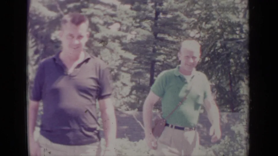 Close 1964 Greenwood Lake home-movie view of two men in polo shirts walking past trees in bright outdoor light.