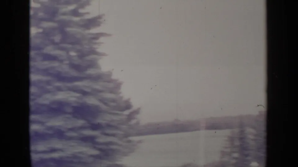 1964 Greenwood Lake home-movie frame showing snowy evergreens, a pale lake surface, distant shoreline, and dark vertical framing.